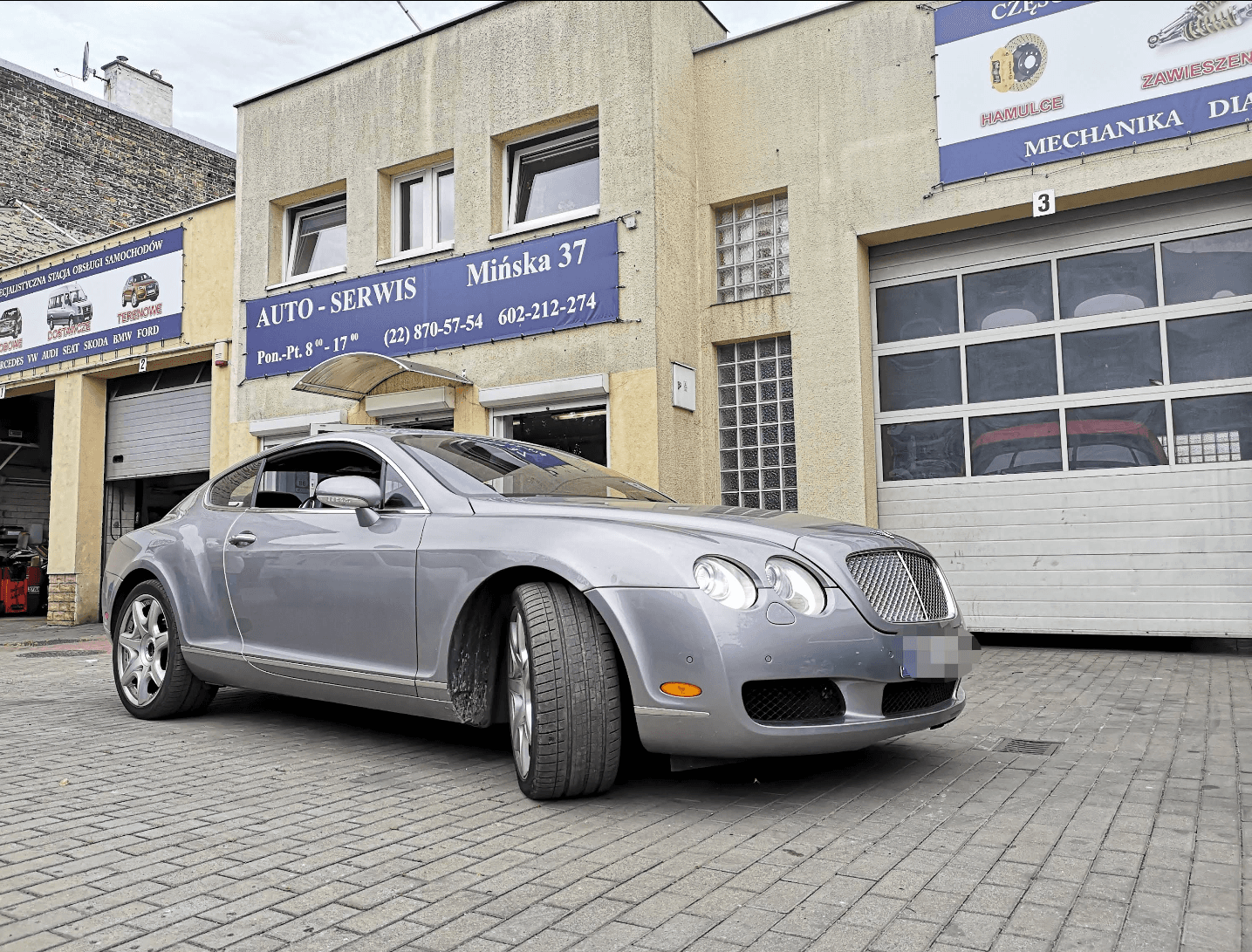 Warsztat Auto-Serwis przy Mińskiej 37 w Warszawie — szary Bentley Continental zaparkowany pod szyldem z godzinami otwarcia i numerami telefonów.